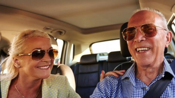 Two older adults sitting in the front seats of the car, smiling with sunglasses on. 