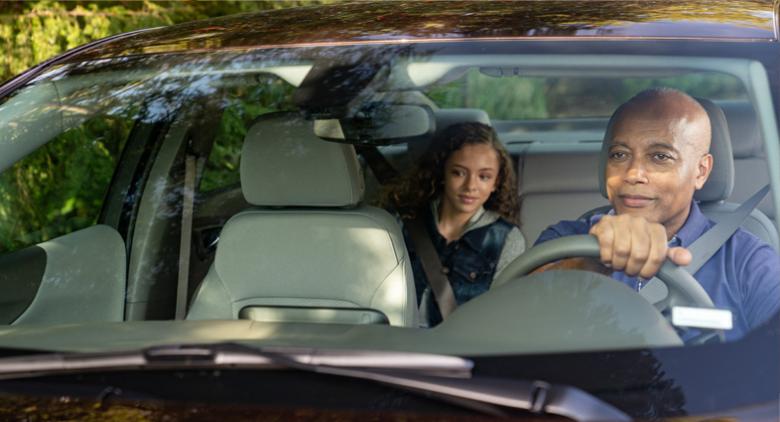 A view through the windshield of an older driver with hands on wheel and seatbelt on, a grandchild is in the backseat also wearing a seatbelt.