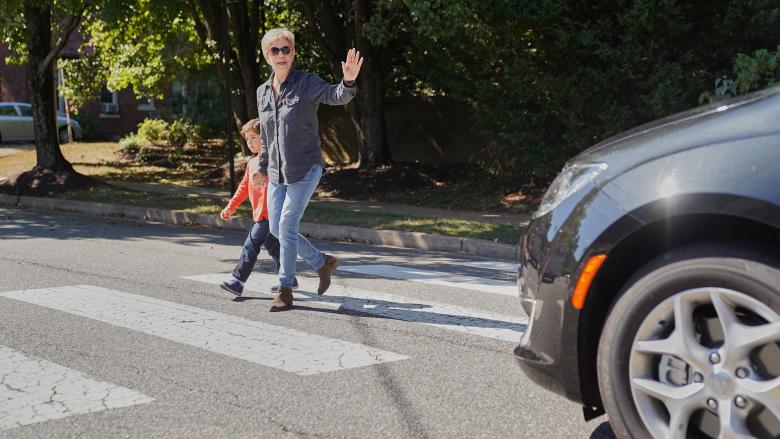 An older adult walks across a crosswalk hand-in-hand with a child, waving at a stopped car whose bumper is in view.