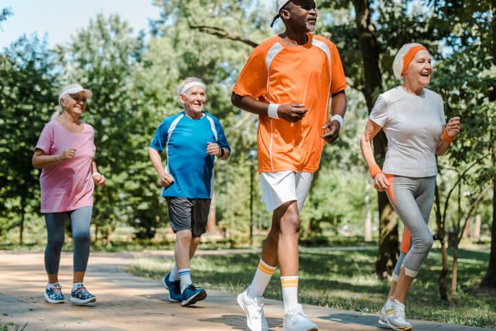 Four older adults in brightly colored athletic-wear jog along a tree-lined path on a sunny Summer day.