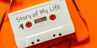 An image of a white cassette tape with "Story of my life" written on the tape. The tape is sitting on an orange table. 