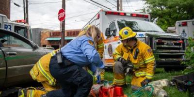 Three emergency medical and rescue services personnel work on a patient involved in a car crash with an ambulance in the background.