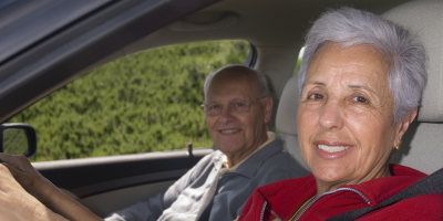 An older woman sitting in the driver seat, smiling toward the camera. A smiling older male is sitting in the passenger seat.