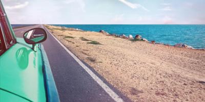 A closeup of a passenger side rearview mirror of a vintage seafoam color car driving along a coastal road with the ocean in the background.